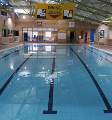 Indoor swimming pool with marked lanes, clear blue water and poolside safety signage, housed within a brick-lined leisure facility with a high roof and skylights.