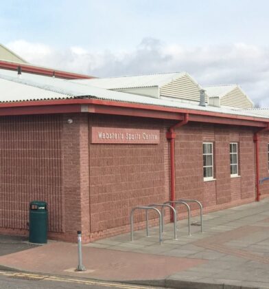 Exterior view of Webster’s Sports Centre, a red-brick leisure facility with blue entrance doors, accessible pathways and bicycle stands outside the main entrance.