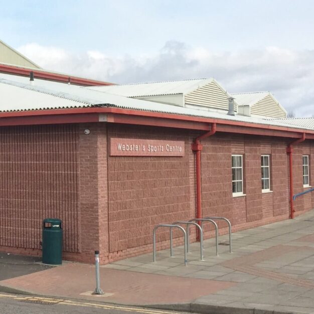 Exterior view of Webster’s Sports Centre, a red-brick leisure facility with blue entrance doors, accessible pathways and bicycle stands outside the main entrance.