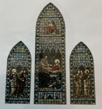 Three Gothic-style stained-glass panels depicting biblical scenes and figures, with intricate architectural details, decorative borders and richly coloured red, blue and gold glass set against a plain wall.
