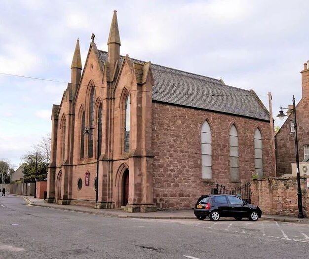 Historic red sandstone church building with Gothic Revival architecture, featuring pointed-arch windows, decorative spires and a steep slate roof, standing on a street corner with neighbouring buildings nearby.