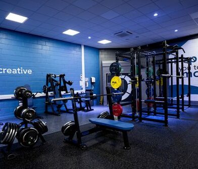 Modern gym with free weights, resistance machines and strength-training equipment arranged across a spacious workout area, featuring blue walls, mirrored surfaces and functional training stations.