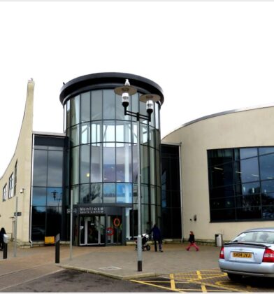 Modern leisure centre building with a distinctive curved façade and glass entrance tower, viewed from the front with pedestrians, parked cars and surrounding walkways.