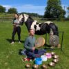 Person seated on grass playing a frame drum and surrounded by singing bowls and a gong, while another person stands beside a black-and-white horse in a fenced field on a sunny day with trees and countryside in the background.