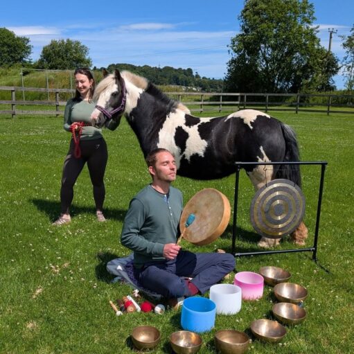 Person seated on grass playing a frame drum and surrounded by singing bowls and a gong, while another person stands beside a black-and-white horse in a fenced field on a sunny day with trees and countryside in the background.