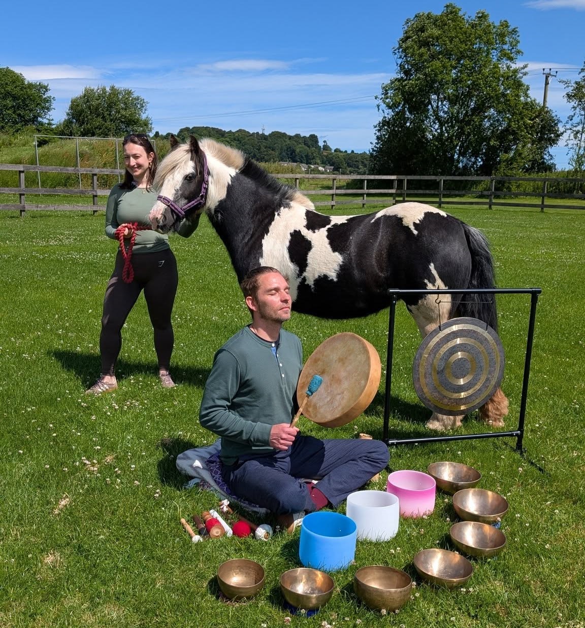 Person seated on grass playing a frame drum and surrounded by singing bowls and a gong, while another person stands beside a black-and-white horse in a fenced field on a sunny day with trees and countryside in the background.