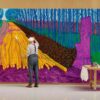 Person standing in a gallery space painting a large, colourful mural depicting a tree-lined avenue and stacks of logs, rendered in vivid purple, blue, green and yellow tones, with an art table and supplies positioned beside the artwork.