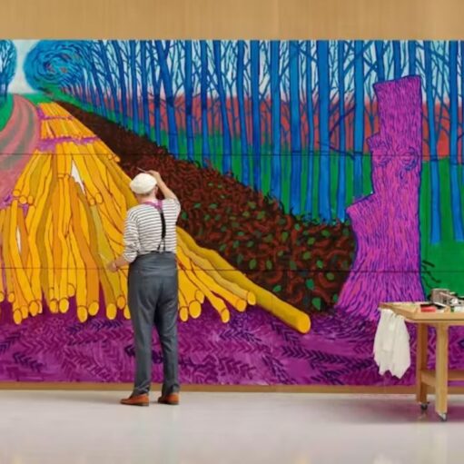 Person standing in a gallery space painting a large, colourful mural depicting a tree-lined avenue and stacks of logs, rendered in vivid purple, blue, green and yellow tones, with an art table and supplies positioned beside the artwork.
