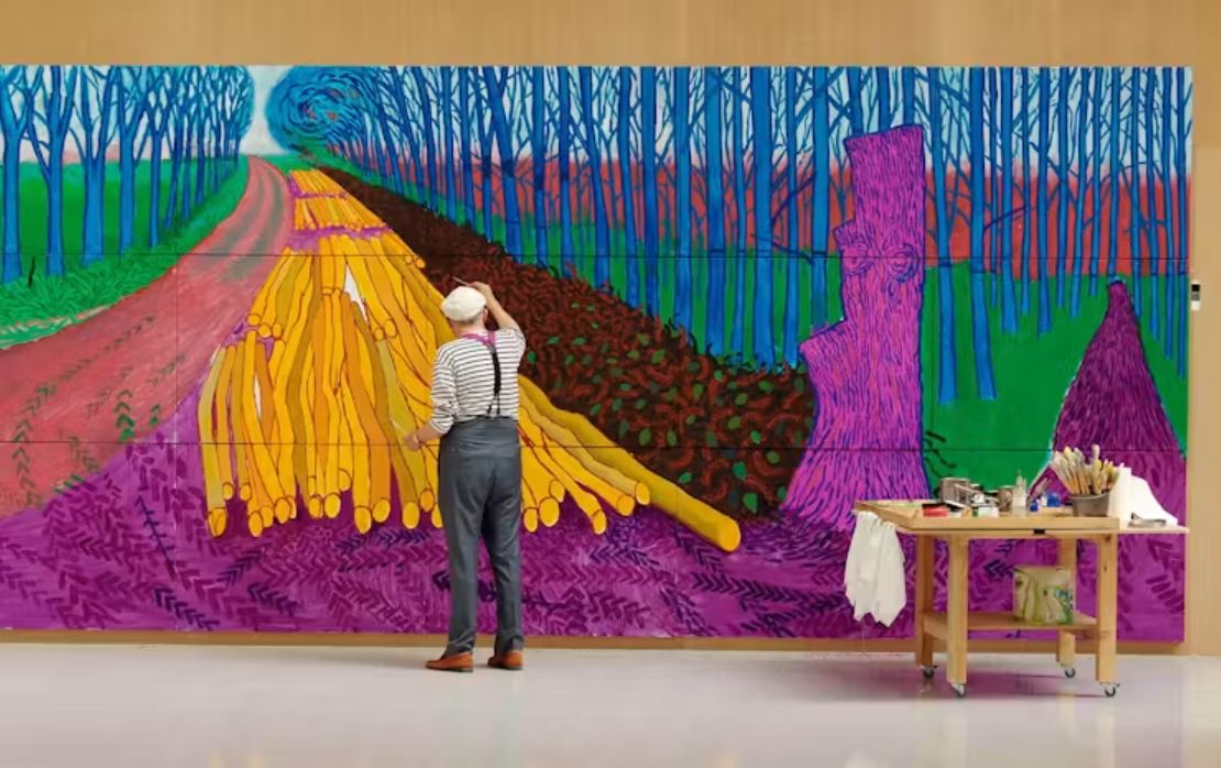 Person standing in a gallery space painting a large, colourful mural depicting a tree-lined avenue and stacks of logs, rendered in vivid purple, blue, green and yellow tones, with an art table and supplies positioned beside the artwork.