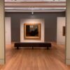 Art gallery interior featuring framed paintings displayed on freestanding walls in a spacious exhibition room, with polished wooden floors, a central bench for visitors and soft gallery lighting highlighting the artwork.