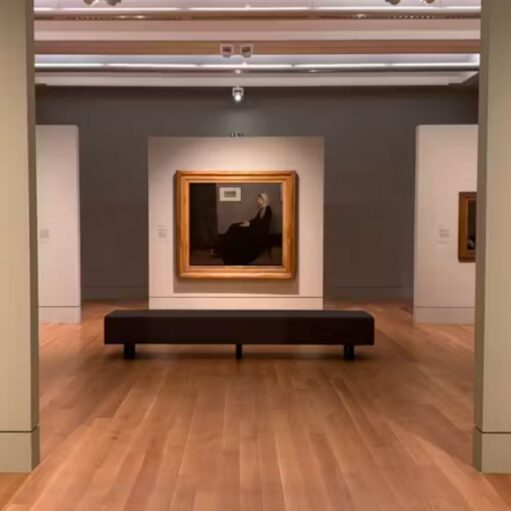 Art gallery interior featuring framed paintings displayed on freestanding walls in a spacious exhibition room, with polished wooden floors, a central bench for visitors and soft gallery lighting highlighting the artwork.