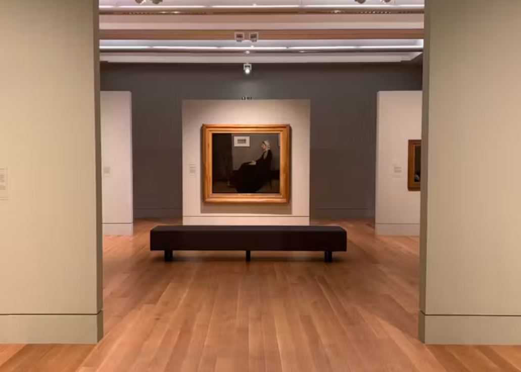 Art gallery interior featuring framed paintings displayed on freestanding walls in a spacious exhibition room, with polished wooden floors, a central bench for visitors and soft gallery lighting highlighting the artwork.
