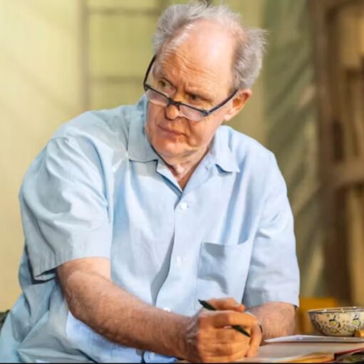 Person wearing glasses and a light blue shirt sits at a table holding a pen, with an open notebook, bowl and drinking glass in front of them, in what appears to be a theatre or stage performance setting with softly lit scenery in the background.