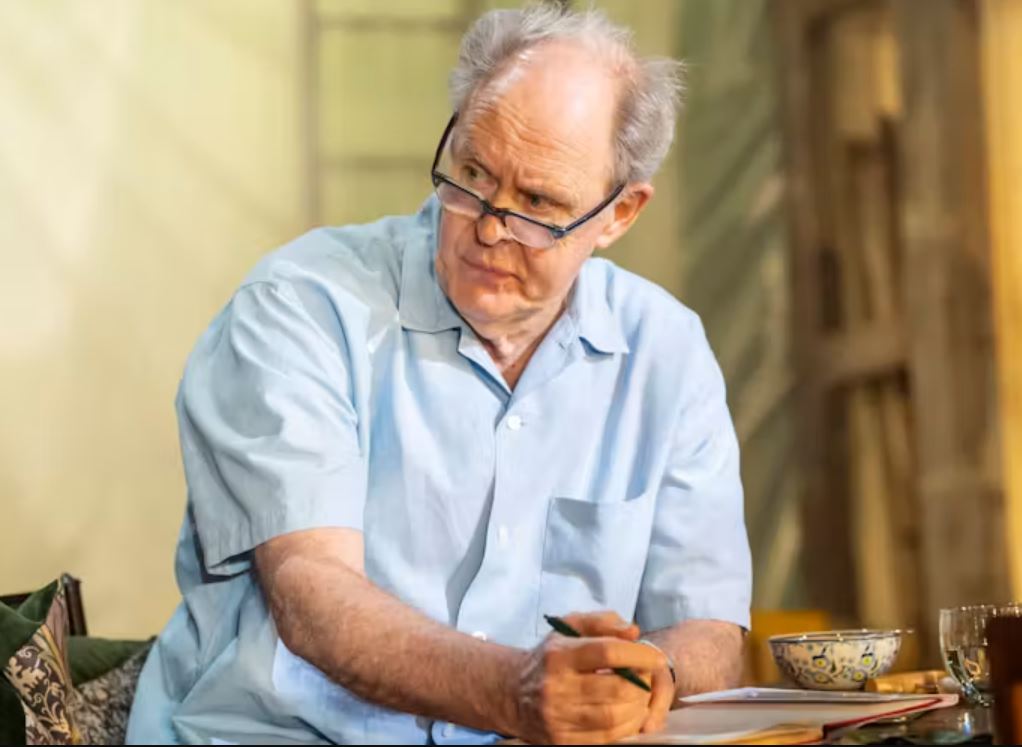 Person wearing glasses and a light blue shirt sits at a table holding a pen, with an open notebook, bowl and drinking glass in front of them, in what appears to be a theatre or stage performance setting with softly lit scenery in the background.