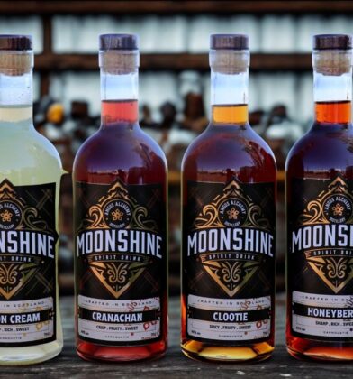 Four bottles of Angus Alchemy Distillery Moonshine spirit drinks displayed on a wooden surface, featuring Lemon Cream, Cranachan, Clootie and Honeyberry flavours, with distinctive black-and-gold labels and varying spirit colours ranging from pale cream to deep amber.