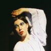Portrait of a person with short dark hair wearing a flowing pale lavender blouse, posed with one arm raised above the head against a dark background, illuminated by dramatic stage-style lighting and wearing bold blue eye makeup.
