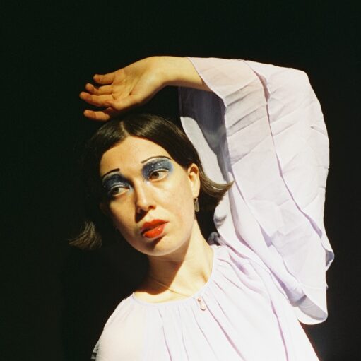 Portrait of a person with short dark hair wearing a flowing pale lavender blouse, posed with one arm raised above the head against a dark background, illuminated by dramatic stage-style lighting and wearing bold blue eye makeup.