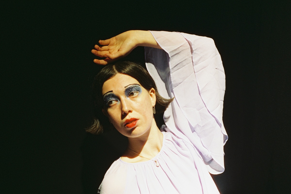 Portrait of a person with short dark hair wearing a flowing pale lavender blouse, posed with one arm raised above the head against a dark background, illuminated by dramatic stage-style lighting and wearing bold blue eye makeup.