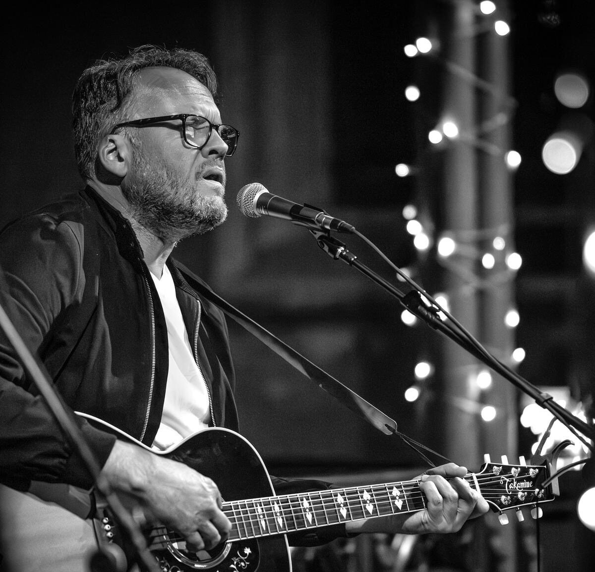 Black-and-white photograph of a musician playing an acoustic guitar and singing into a microphone on stage, with decorative string lights blurred in the background and concert equipment visible in the foreground.