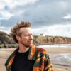 Portrait of a person standing on a sandy beach wearing a colourful patterned jacket, with rugged cliffs, shoreline and sea in the background beneath a dramatic cloudy sky.