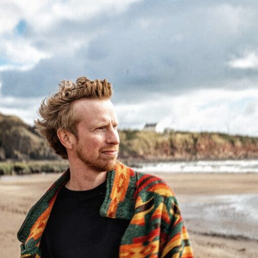 Portrait of a person standing on a sandy beach wearing a colourful patterned jacket, with rugged cliffs, shoreline and sea in the background beneath a dramatic cloudy sky.