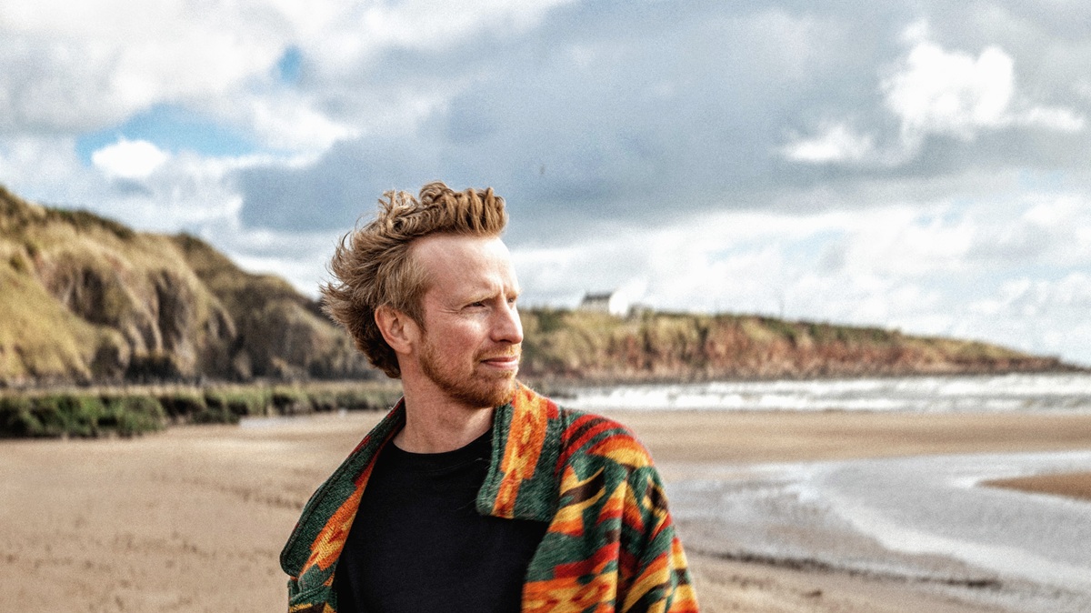 Portrait of a person standing on a sandy beach wearing a colourful patterned jacket, with rugged cliffs, shoreline and sea in the background beneath a dramatic cloudy sky.