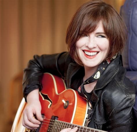 Person seated with a hollow-body electric guitar, wearing a black leather jacket and patterned shirt, looking toward the camera in an indoor setting with a warm-toned background.