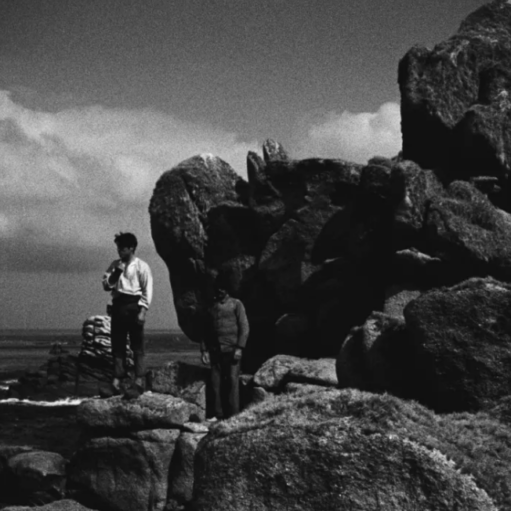 Black-and-white still image showing two people standing on a rocky coastal outcrop beside large granite boulders, overlooking the sea under a dramatic sky with dense clouds.
