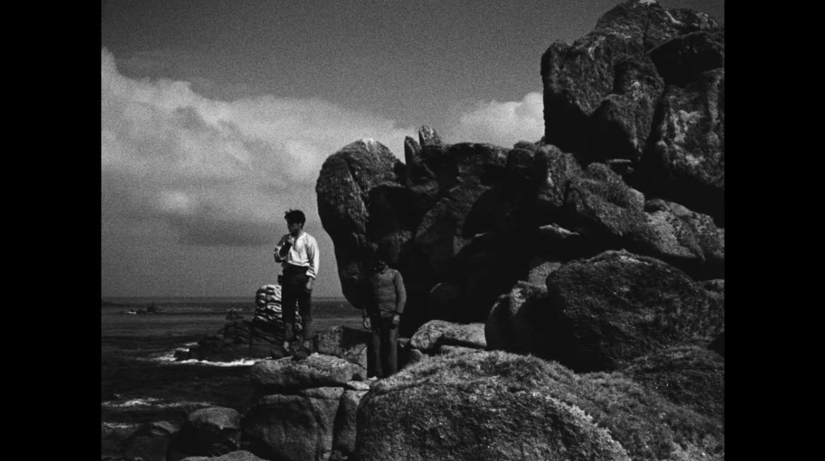 Black-and-white still image showing two people standing on a rocky coastal outcrop beside large granite boulders, overlooking the sea under a dramatic sky with dense clouds.