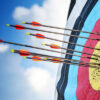 Close-up of an archery target outdoors, with multiple arrows clustered around the yellow bullseye and inner scoring rings, set against a bright blue sky with scattered clouds.