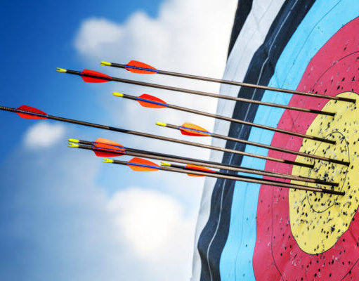 Close-up of an archery target outdoors, with multiple arrows clustered around the yellow bullseye and inner scoring rings, set against a bright blue sky with scattered clouds.