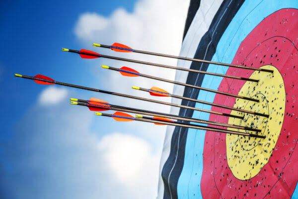 Close-up of an archery target outdoors, with multiple arrows clustered around the yellow bullseye and inner scoring rings, set against a bright blue sky with scattered clouds.
