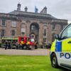 Police Scotland vehicles and a fire engine parked in front of a large historic stone building during a community or emergency services event, with visitors gathered in the courtyard and a Scottish flag flying above the entrance.