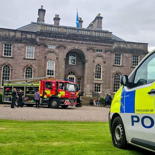 Police Scotland vehicles and a fire engine parked in front of a large historic stone building during a community or emergency services event, with visitors gathered in the courtyard and a Scottish flag flying above the entrance.