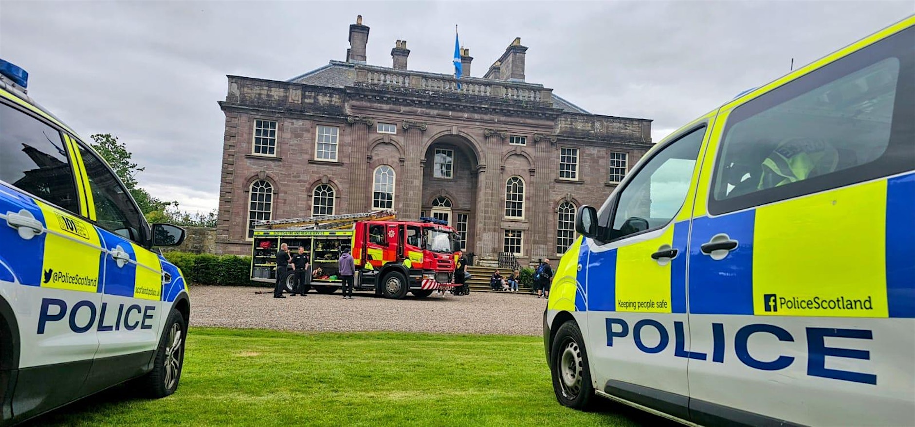 Police Scotland vehicles and a fire engine parked in front of a large historic stone building during a community or emergency services event, with visitors gathered in the courtyard and a Scottish flag flying above the entrance.