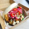 Steamed bao bun filled with crispy coated chicken, topped with pickled red onions, sliced red chillies, spring onions and sesame seeds, drizzled with creamy mayonnaise and dark sauce, served in a cardboard takeaway tray.