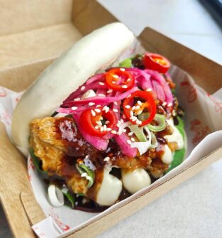 Steamed bao bun filled with crispy coated chicken, topped with pickled red onions, sliced red chillies, spring onions and sesame seeds, drizzled with creamy mayonnaise and dark sauce, served in a cardboard takeaway tray.