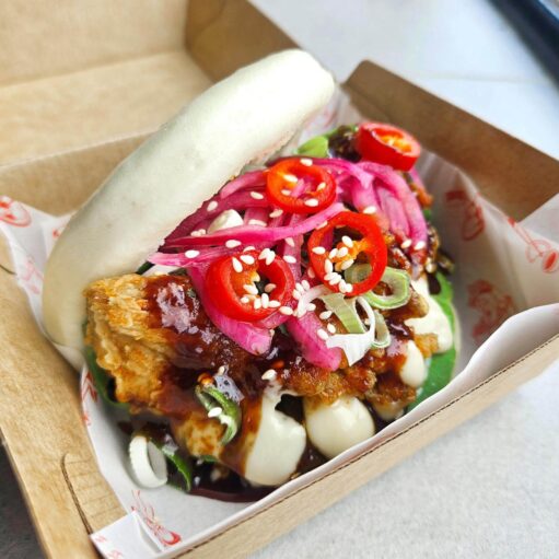 Steamed bao bun filled with crispy coated chicken, topped with pickled red onions, sliced red chillies, spring onions and sesame seeds, drizzled with creamy mayonnaise and dark sauce, served in a cardboard takeaway tray.
