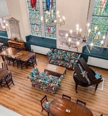 Interior of a café housed within a converted church, featuring stained-glass windows, wooden tables and chairs, floral-patterned sofas, chandeliers and a grand piano, viewed from an elevated balcony overlooking the bright open space.