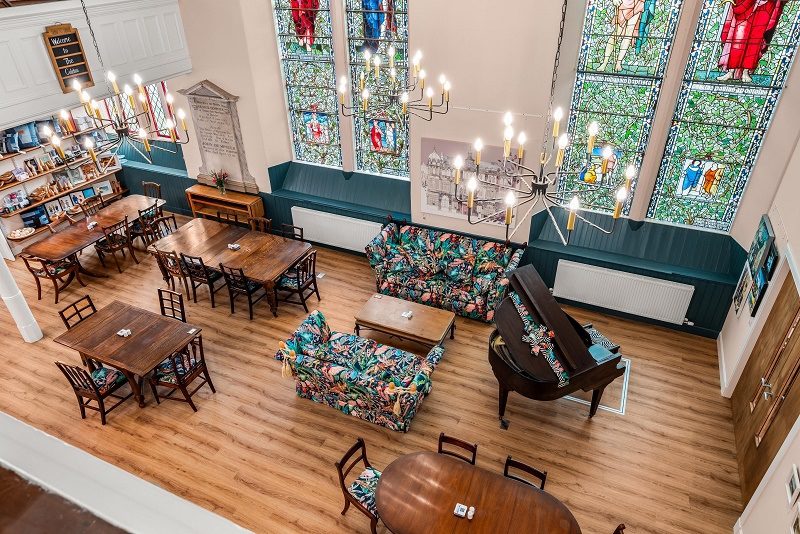 Interior of a café housed within a converted church, featuring stained-glass windows, wooden tables and chairs, floral-patterned sofas, chandeliers and a grand piano, viewed from an elevated balcony overlooking the bright open space.