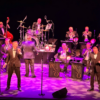 Live big band performance on a theatre stage featuring a large jazz orchestra seated behind black music stands marked “MSO”. Two vocalists stand at the front of the stage with microphones, while a conductor faces the orchestra from the centre. The ensemble includes brass players, saxophonists, a drummer, and a double bass player arranged across several rows. Purple and pink stage lighting illuminates the performers and floor, while the background remains dark, drawing attention to the musicians and instruments. The image conveys the scale of a live swing or big band concert with a full orchestra and featured singers performing together.