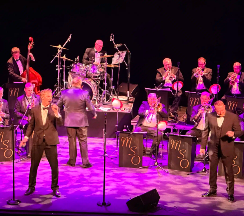 Live big band performance on a theatre stage featuring a large jazz orchestra seated behind black music stands marked “MSO”. Two vocalists stand at the front of the stage with microphones, while a conductor faces the orchestra from the centre. The ensemble includes brass players, saxophonists, a drummer, and a double bass player arranged across several rows. Purple and pink stage lighting illuminates the performers and floor, while the background remains dark, drawing attention to the musicians and instruments. The image conveys the scale of a live swing or big band concert with a full orchestra and featured singers performing together.