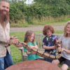 Outdoor wildlife encounter featuring four people gathered around a large patterned snake in a grassy park setting. One person with a long beard and visible arm tattoos stands to the left holding part of the snake, while two children and another adult seated on a wooden bench support the snake's body along its length.
