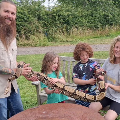Outdoor wildlife encounter featuring four people gathered around a large patterned snake in a grassy park setting. One person with a long beard and visible arm tattoos stands to the left holding part of the snake, while two children and another adult seated on a wooden bench support the snake's body along its length.
