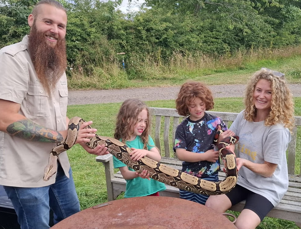 Outdoor wildlife encounter featuring four people gathered around a large patterned snake in a grassy park setting. One person with a long beard and visible arm tattoos stands to the left holding part of the snake, while two children and another adult seated on a wooden bench support the snake's body along its length.