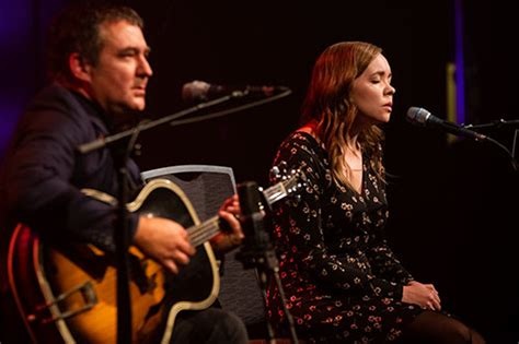 Live acoustic music performance on a theatre stage featuring two seated performers positioned at separate microphones. One performer is playing an acoustic guitar while seated to the left of the stage, and the other performer is seated to the right singing into a microphone. The stage is lit with warm red and purple lighting against a dark background, drawing attention to the performers.