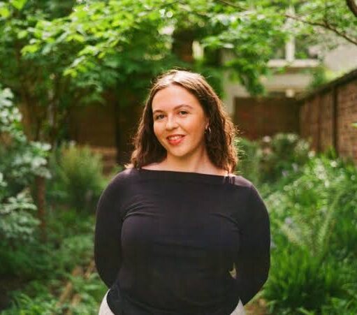 Portrait photograph of a person standing in a lush garden surrounded by dense green foliage, shrubs and trees. The person is positioned centrally on a narrow garden path, wearing a plain dark long-sleeved top and light-coloured trousers. The setting features layered planting with a variety of leafy textures and shades of green, creating a natural and tranquil outdoor environment.