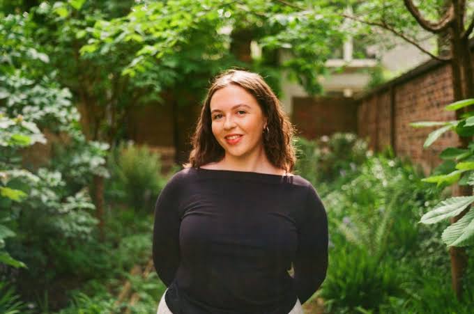 Portrait photograph of a person standing in a lush garden surrounded by dense green foliage, shrubs and trees. The person is positioned centrally on a narrow garden path, wearing a plain dark long-sleeved top and light-coloured trousers. The setting features layered planting with a variety of leafy textures and shades of green, creating a natural and tranquil outdoor environment.