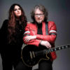 Promotional photograph featuring two musicians posed against a plain dark grey studio background. One person stands on the left wearing dark clothing, while another stands on the right wearing a bright red leather jacket and resting folded arms on a black electric guitar. The guitar is positioned diagonally across the foreground, with its neck extending toward the right edge of the image.
