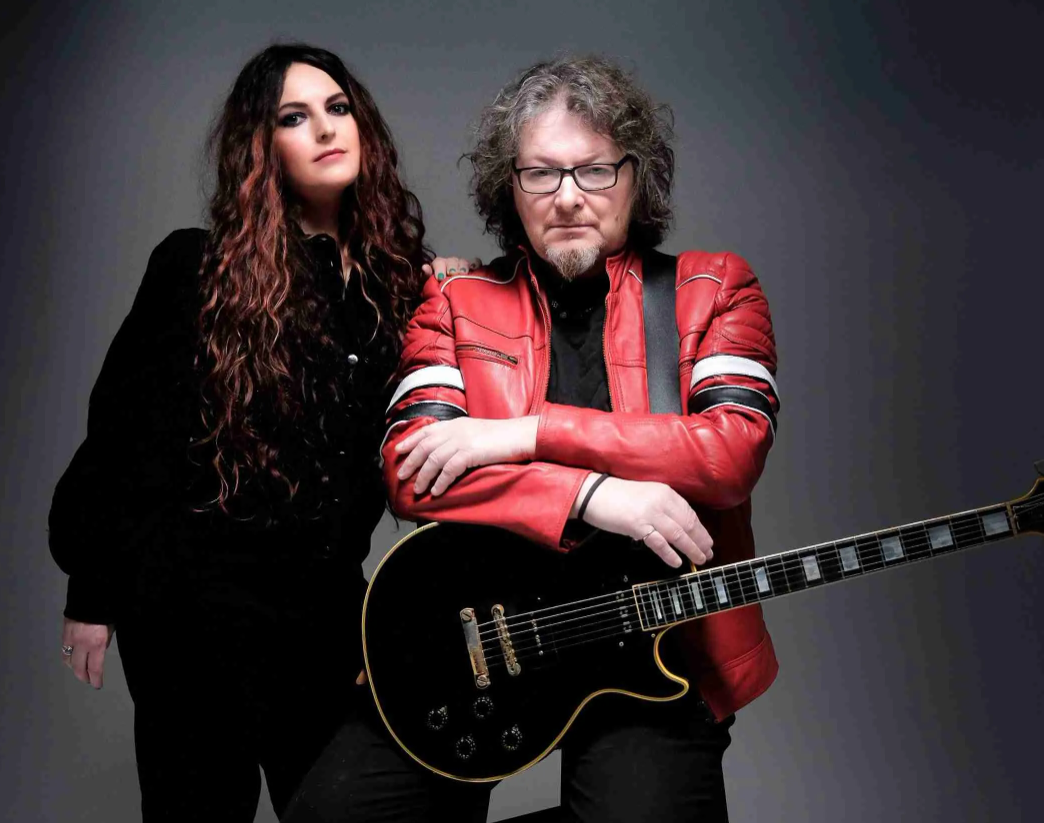 Promotional photograph featuring two musicians posed against a plain dark grey studio background. One person stands on the left wearing dark clothing, while another stands on the right wearing a bright red leather jacket and resting folded arms on a black electric guitar. The guitar is positioned diagonally across the foreground, with its neck extending toward the right edge of the image.
